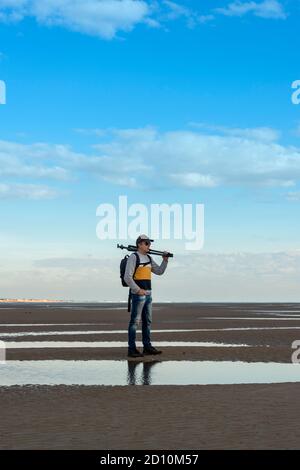 Uomo che porta un treppiede in piedi sulla sabbia presso la costa. Fotografo con attrezzatura. Foto Stock