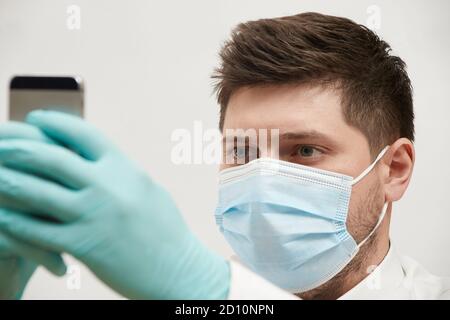 concetto di coronavirus un uomo in una maschera medica guarda il telefono si trova su uno sfondo bianco Foto Stock