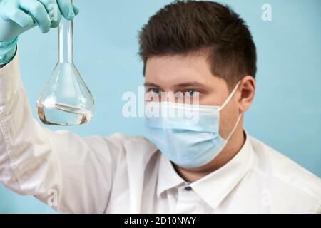 l'uomo di concetto del coronavirus in maschera medica guarda la bottiglia sopra sfondo blu Foto Stock