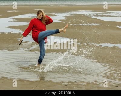 Femmina sulla spiaggia che calcia l'acqua in piedi in una pozza Foto Stock