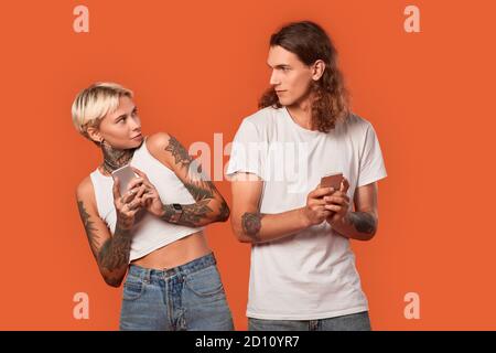 Stile libero. Giovane coppia tatuata in piedi isolati in arancione con smartphone che si guardano l'un l'altro informazioni nervose nascondendo Foto Stock