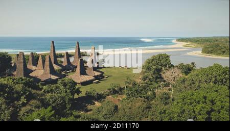 Vista aerea del villaggio tradizionale con case ornately tetto sulla spiaggia di sabbia spiaggia di mare con alberi tropici e piante. Valle verde erba sulla costa sabbiosa oceano Isola di Sumba, Indonesia, Asia a colpo drone Foto Stock