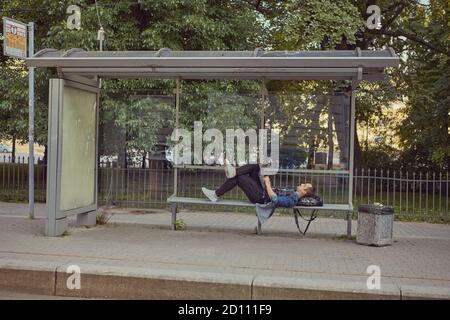 Un uomo bianco alla fermata dell'autobus si stese su una panchina con un computer portatile in ginocchio. Un giovane maschio usa un computer portatile in attesa dei trasporti pubblici. Foto Stock
