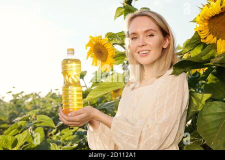 Giovane donna bionda sorridente in abito bianco che tiene una bottiglia di olio di girasole Foto Stock