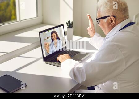 Medico senior in uniforme e occhiali seduti al computer portatile e. saluto giovane donna sorridente paziente online Foto Stock