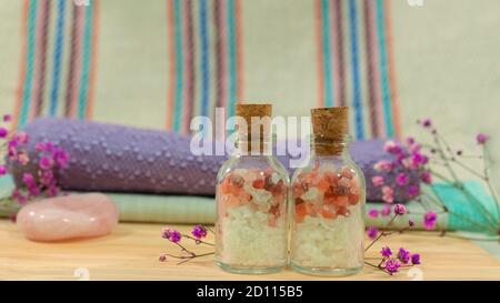 Composizione con due bottiglie di vetro con sali da bagno su tavola di legno con asciugamani, pietra rosa, fiori viola e panno con linee sullo sfondo Foto Stock