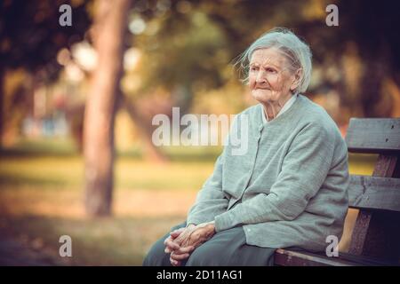 Ritratto di donna anziana seduta su panchina nel parco autunnale. Vecchia signora sentendosi sola e triste. Frustrata femmina anziano all'aperto. Foto Stock