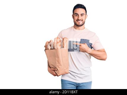 Giovane bell'uomo che tiene il sacchetto del pane sorridente che punta felice con mano e dito Foto Stock