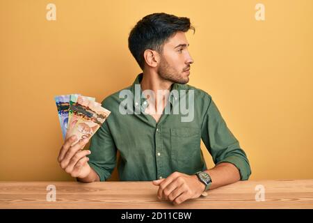 Giovane bell'uomo che tiene dollari canadesi guardando a lato, rilassa il profilo posa con viso naturale e sorriso fiducioso. Foto Stock