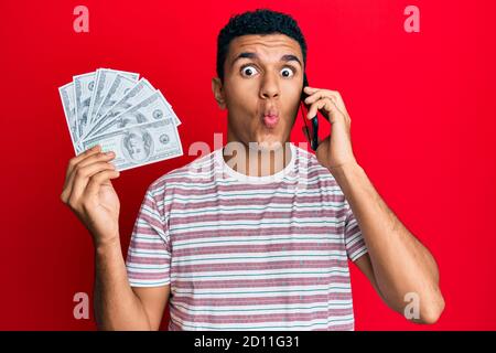 Giovane arabo che ha conversazione parlando sullo smartphone che tiene dollari facendo il pesce faccia con la bocca e squinting occhi, pazzo e comico. Foto Stock