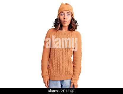 Giovane bella donna mista da corsa con maglione di lana e cappello invernale che fa viso di pesce con labbra, gesto pazzo e comico. Espressione divertente. Foto Stock