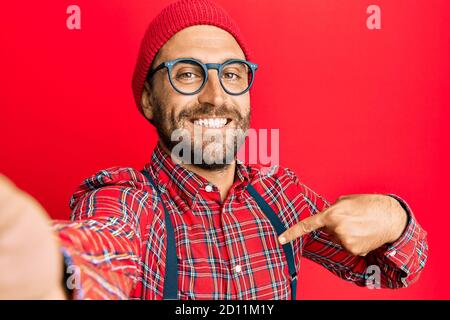 Bell'uomo hipster con barba scattando una foto selfie con smartphone che punta il dito verso un sorriso di sé felice e orgoglioso Foto Stock