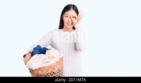 Giovane bella donna cinese che tiene cesto di bucato con vestiti sorridente buon fare ok segno con mano su occhio guardando attraverso dita Foto Stock