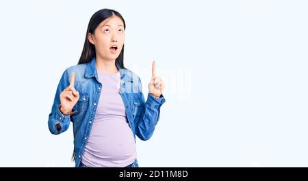 Giovane bella donna cinese incinta attesa bambino stupito e sorpreso guardando in su e puntando con le dita e le braccia sollevate. Foto Stock