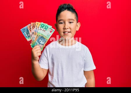 Bambino ispanico ragazzo piccolo che tiene dollari australiani che sembrano positivi e. felice in piedi e sorridente con un sorriso sicuro che mostra i denti Foto Stock