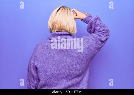 Bella donna bionda Plus taglia indossa un pullover casual con dolcevita Sfondo viola all'indietro pensando al dubbio con la mano sulla testa Foto Stock