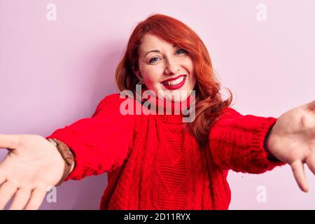 Giovane bella donna rossa che indossa un pullover rosso casual turtleneck su sfondo rosa guardando la fotocamera sorridente con le braccia aperte per abbraccio. Cheerfu Foto Stock