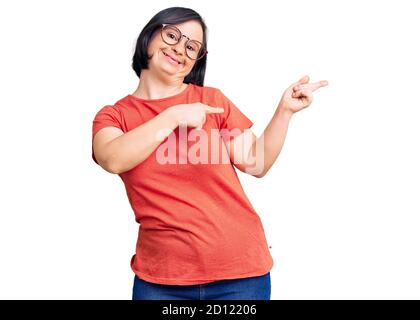 Donna bruna con sindrome di Down indossare abiti e occhiali casual sorridendo e guardando la fotocamera con due mani e le dita di lato Foto Stock