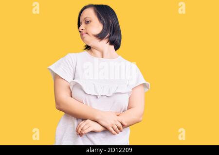 Donna bruna con sindrome di Down indossare casual tshirt bianca cercando al fianco con le braccia incrociate convinte e fiduciose Foto Stock