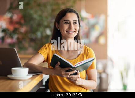 Giovane donna entusiasta seduto in un bar, usando un computer portatile, tenendo un blocco note Foto Stock