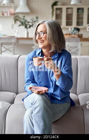 Felice rilassata donna anziana matura bere caffè rilassante sul divano a casa. Foto Stock