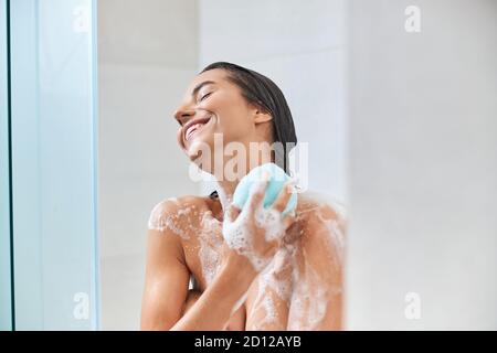 Giovane donna gioiosa che usa la loofah del bagno mentre prende la doccia Foto Stock