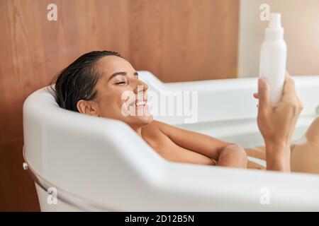 Giovane donna di charme che tiene una bottiglia di lozione mentre si prende il bagno Foto Stock