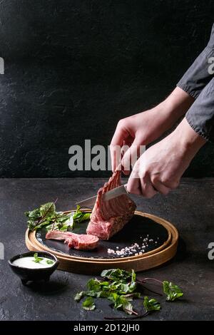 Uomo con le mani in mano il taglio delle grigliate di carré di agnello con yogurt salsa alla menta servito con insalata verde giovani foglie di bietole e sale rosa sul round ardesia in legno Foto Stock