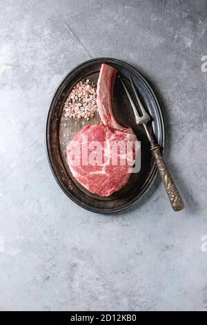 Bistecca di tomahawk nero crudo di angus, non cotta, servita con sale, pepe e forchetta di carne su vassoio di metallo vintage su fondo grigio. Vi Foto Stock