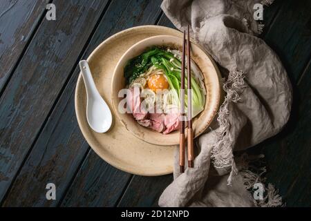 Piatto asiatico udon tagliatelle con tuorlo d'uovo, sesamo, funghi, boc choy, affettato sous vide carne cotta servito in tazza ceramica con cucchiaio e bastoncini su c Foto Stock
