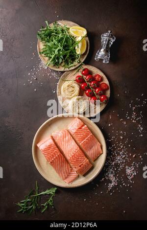 Fette di materie non cotte di filetto di salmone su piastra in ceramica con gli ingredienti per la cena, rucola, limone, sale, pepe, tagliatelle, pomodori ciliegia oltre il marrone scuro Foto Stock