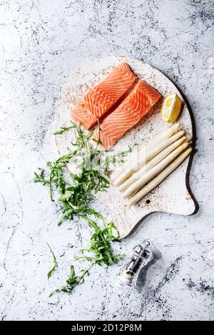 Fette di materie non cotte di filetto di salmone sul tagliere di legno a forma di pesce con gli ingredienti per cucinare la cena rucola, asparago bianco su bianco marbl Foto Stock