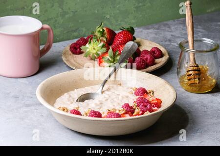 Riso dolce budino di porridge in piastra di ceramica con frutti di bosco fragola e lampone, noci, miele e la tazza di latte su grigio tavolo da cucina con verde wa Foto Stock