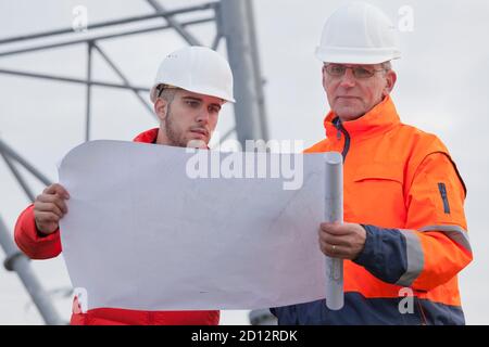 Architetto giovane e maturo con elmetto che guarda un piano di costruzione in un cantiere o piattaforma petrolifera - focalizzazione selettiva sulle persone Foto Stock