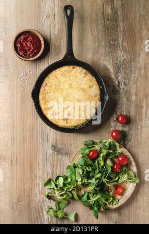 Lo spagnolo frittata di patate tortilla con pancetta servita in ghisa padella con il sugo di pomodoro e gli ingredienti sopra lo sfondo di legno. Vista superiore, spazio. Foto Stock