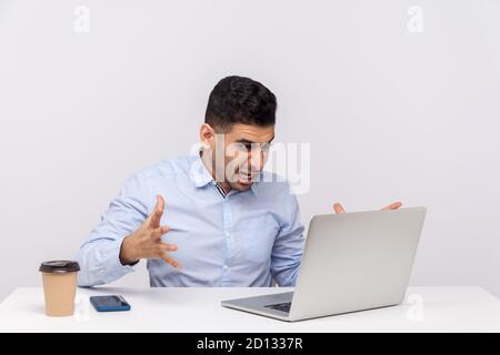 Irritato uomo d'affari seduto in ufficio, guardando un portatile con un'espressione furiosa, gridando arrabbiato mentre si ha conversazione online, videochiamata Foto Stock