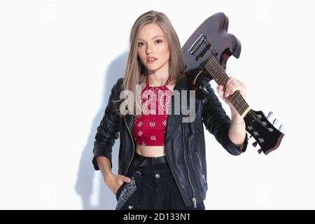 Musicista motivato che tiene la mano in tasca e la chitarra sulla spalla, indossando una giacca in pelle mentre si sta in piedi su sfondo bianco studio Foto Stock