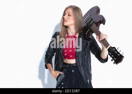 Affascinante musicista che tiene la chitarra a spalla e a mano in tasca, indossando una giacca in pelle mentre si trova su uno sfondo bianco dello studio Foto Stock