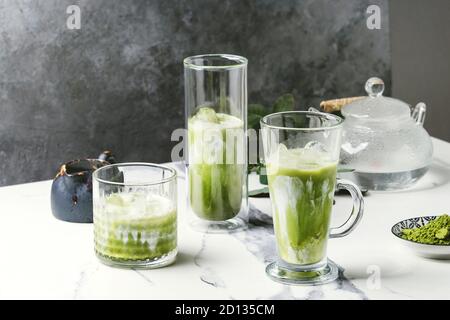 Tè verde Matcha iced latte macchiato o un cocktail in tre diversi tipi di bicchieri con cubetti di ghiaccio, matcha polvere e il contenitore del latte su bianco tavola di marmo, decorata da g Foto Stock