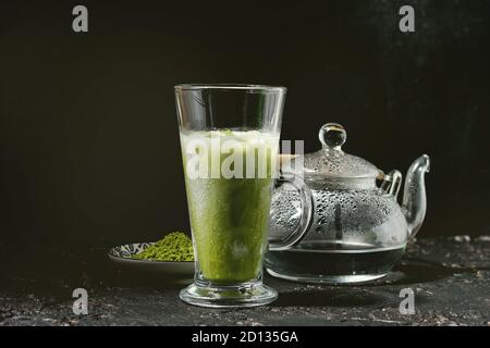 Tè verde Matcha iced latte macchiato o un cocktail nel bicchiere alto con cubetti di ghiaccio e menta, matcha polvere trasparente e teiera con acqua calda sulla texture scuro indietro Foto Stock