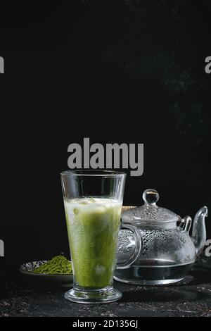 Tè verde Matcha iced latte macchiato o un cocktail nel bicchiere alto con cubetti di ghiaccio e menta, matcha polvere trasparente e teiera con acqua calda sulla texture scuro indietro Foto Stock
