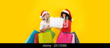Due giovani donne carine che indossano il cappello di Babbo Natale guardando alla macchina fotografica con viso stupefatto, bocca aperta, occhi larghi, tenendo borse colorate per la spesa - Cristo Foto Stock