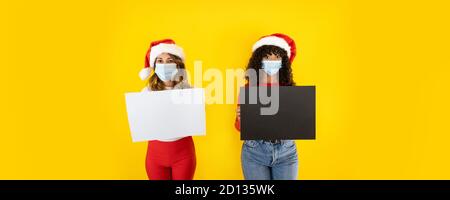 Due giovani donne carine che indossano il cappello di Babbo Natale guardando alla macchina fotografica con gli occhi larghi e la maschera medica, tenendo il segno per lo spazio di copia - banner di Natale per Foto Stock