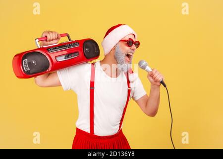 Uomo anziano positivo e attivo nel cappello di babbo natale e occhiali da sole che tengono il registratore a nastro sulla spalla canti al microfono, karaoke di festa. Indo Foto Stock