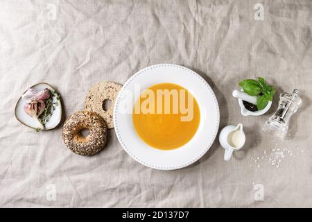 Piastra della zucca vegetariano zuppa di carota servita con erbe aromatiche, cucchiaio, brocca di crema, bagel pane, pepe nero, aglio sopra tela di lino come backg Foto Stock