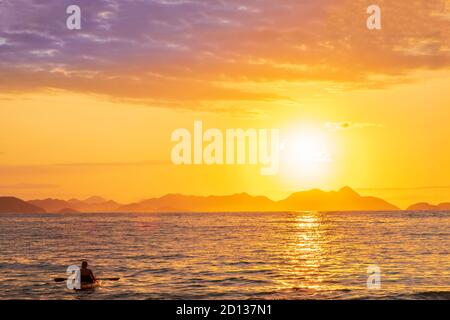 Un canoista all'alba con il sole che sorge su una tranquilla baia tropicale dell'oceano e colline lontane Foto Stock