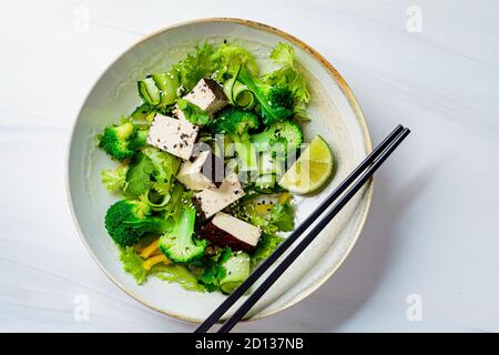 Insalata asiatica verde con broccoli e tofu affumicato in una ciotola bianca, fondo bianco. Foto Stock