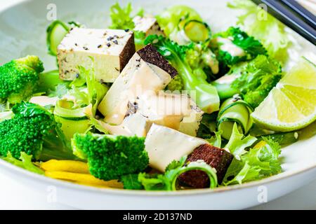 Insalata asiatica verde con broccoli, tofu affumicato e salsa di arachidi in una ciotola bianca, fondo bianco. Foto Stock