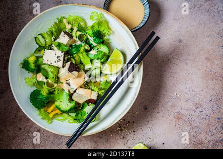Insalata vegana verde con broccoli, tofu affumicato e condimento tahini in una ciotola bianca, fondo scuro. Foto Stock