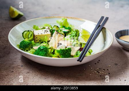 Insalata vegana verde con broccoli, tofu affumicato e condimento tahini in una ciotola bianca, fondo scuro. Foto Stock